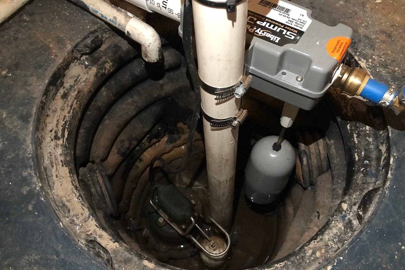 Overhead view of a basement sump basin with a pump, float switch, pump controller, and PVC discharge line